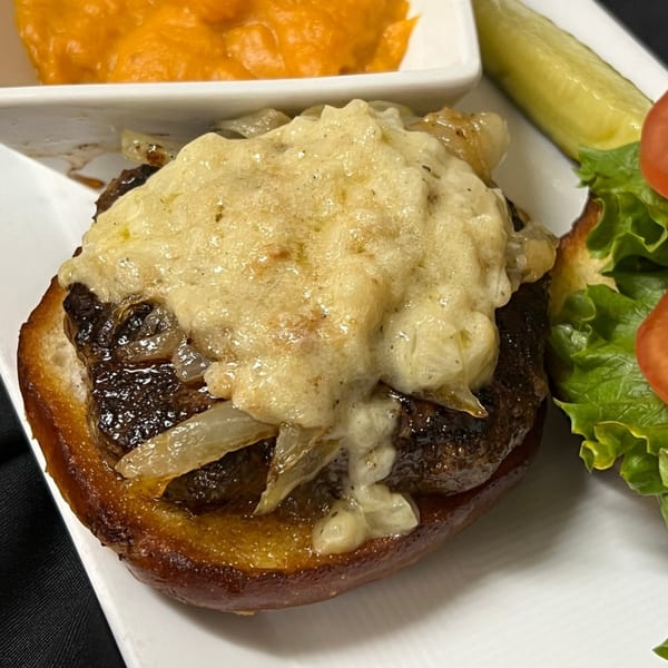 A burger patty topped with melted cheese and caramelized onions, served with a side of sweet potato puree, a pickle, and a small salad.